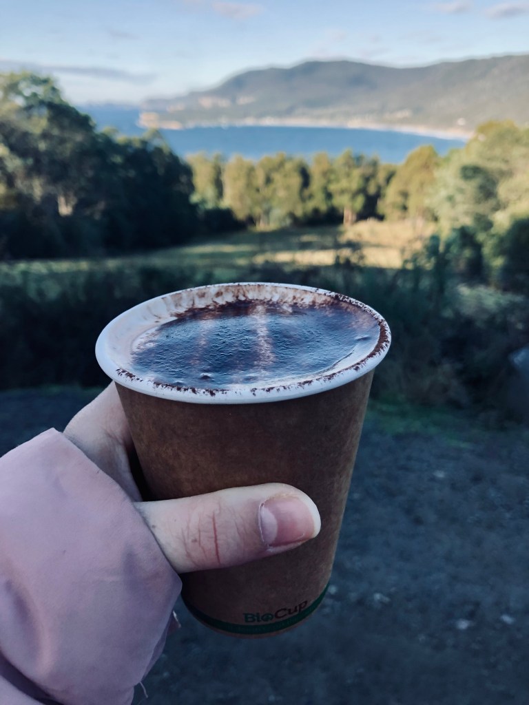 Hand holding hot chocolate with views of ocean and hills in the distance in Tasmania