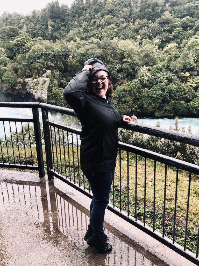 Girl smiling in front of Huka Falls in black rain coat in New Zealand
