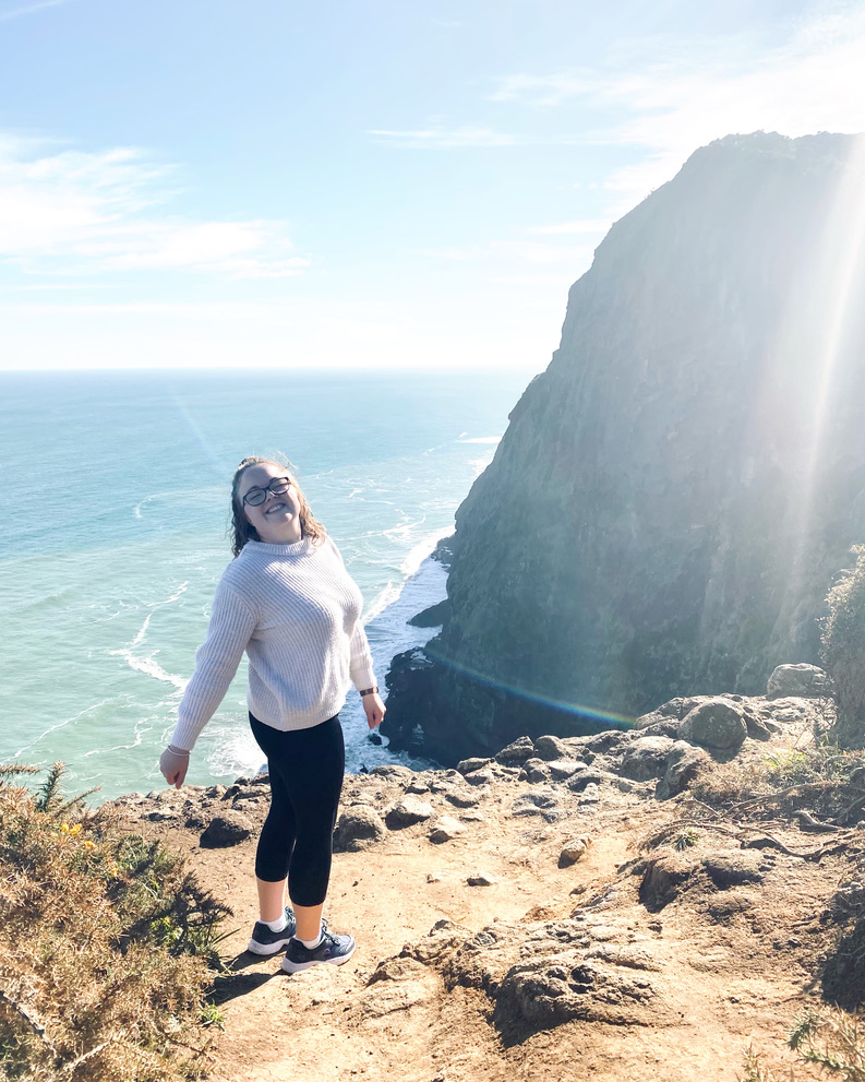 Girl in white sweater smiling in front of views of cliff edge in Mercer Bay Loop in New Zealand's North Island