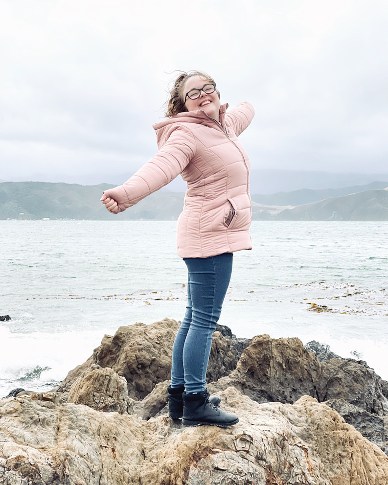 Girl in pink jacket smiling on a rock with ocean in background in Wellington