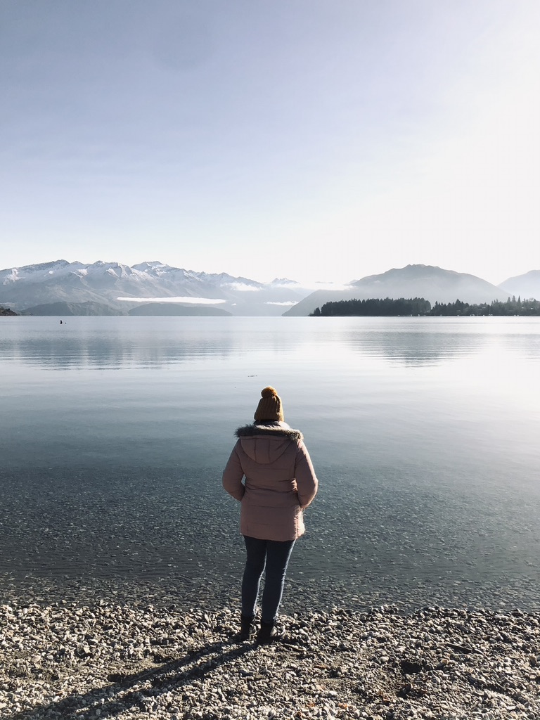 Girl in pink jacket looking out at crystal clear Lake Wanaka with mountains in distance on a sunny day in New Zealand