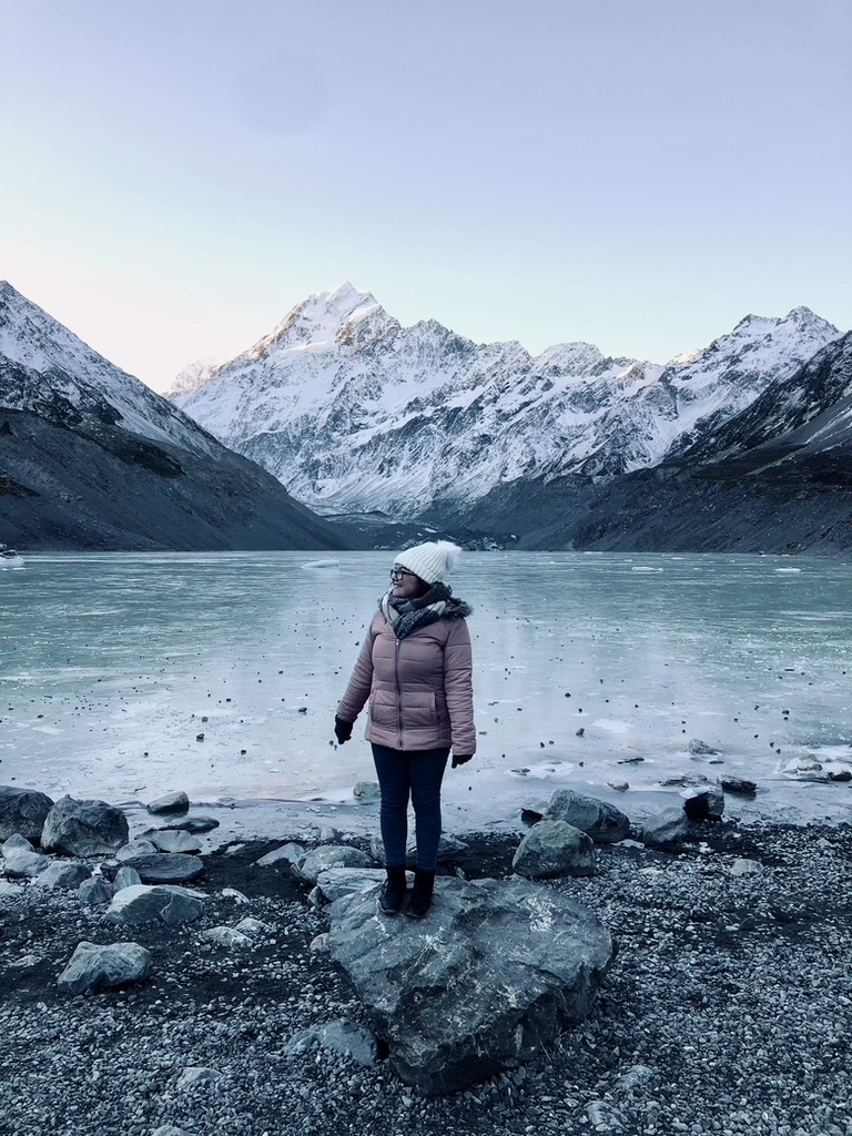 Girl in pink jacket looking to the left on a rock in front of frozen glacier lake with Mt Cook in the background at Mt Cook National Park in New Zealand.