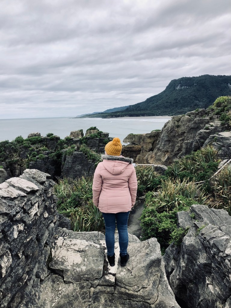 Girl in pink jacket standing on a rock looking out at coastline with ocean in the distance at Pancake Rocks in New Zealand