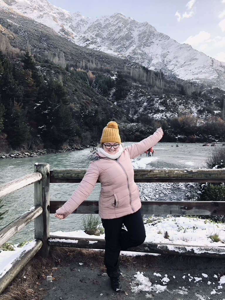 Girl in pink jacket smiling in front of snow capped mountains and river near Queenstown, New Zealand
