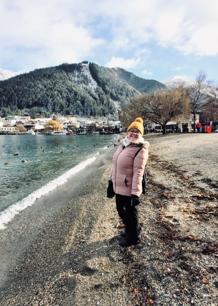 Girl in pink jacket smiling on the sand at lake in Queenstown, New Zealand