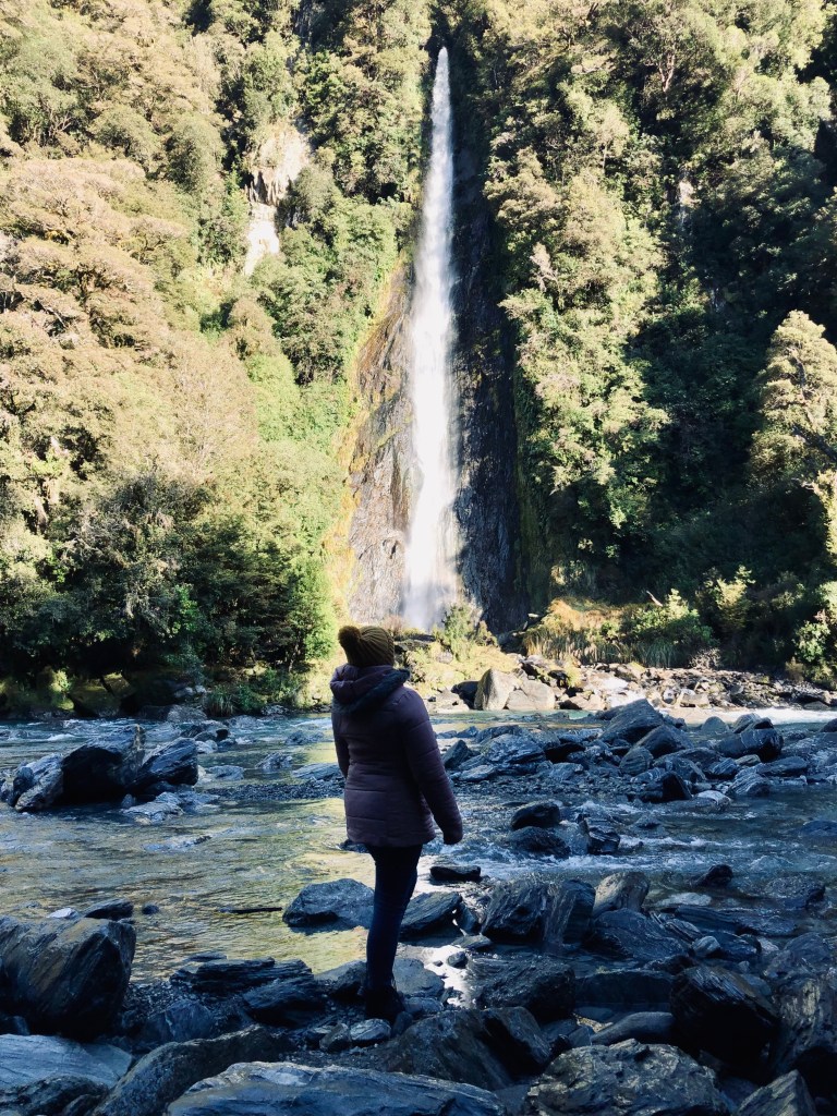 Girl looking up at tall waterfall with green trees surrounding on a road trip on New Zealand's South Island