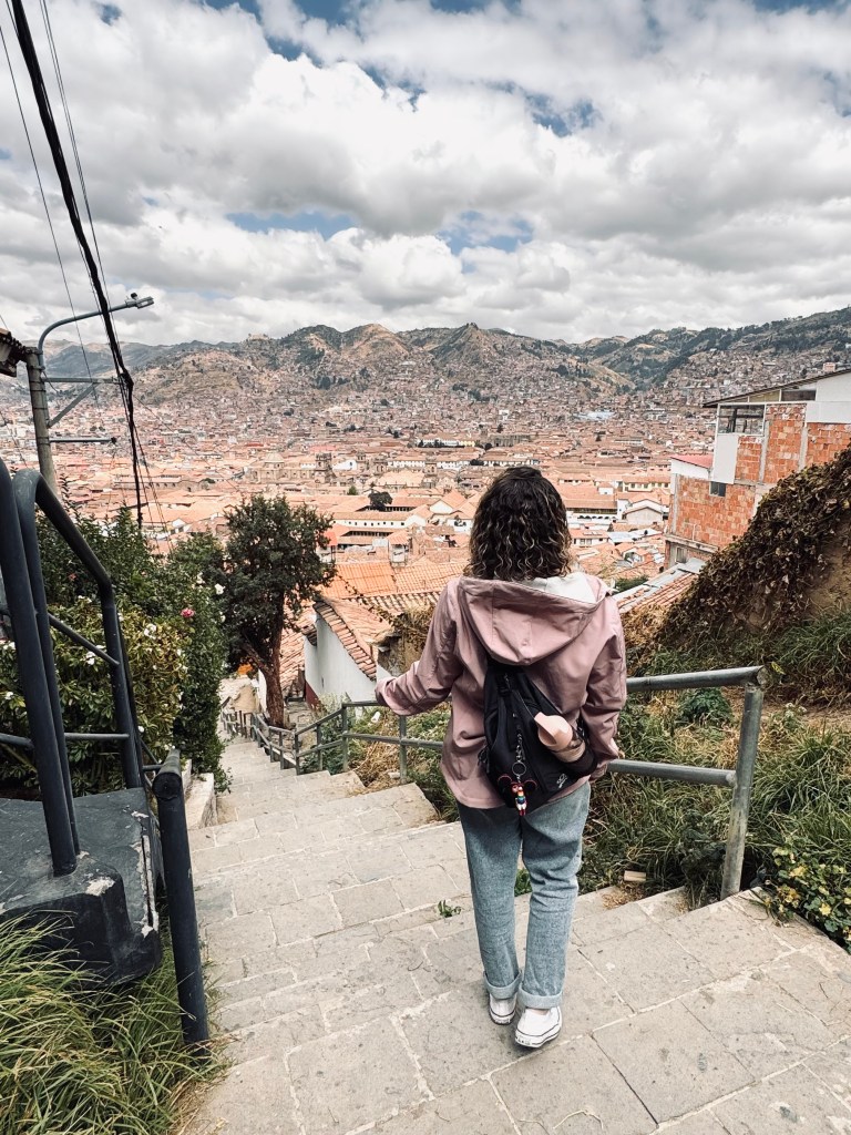 Girl in pink jacket looking out at view of Cusco city with mountains in background in Peru