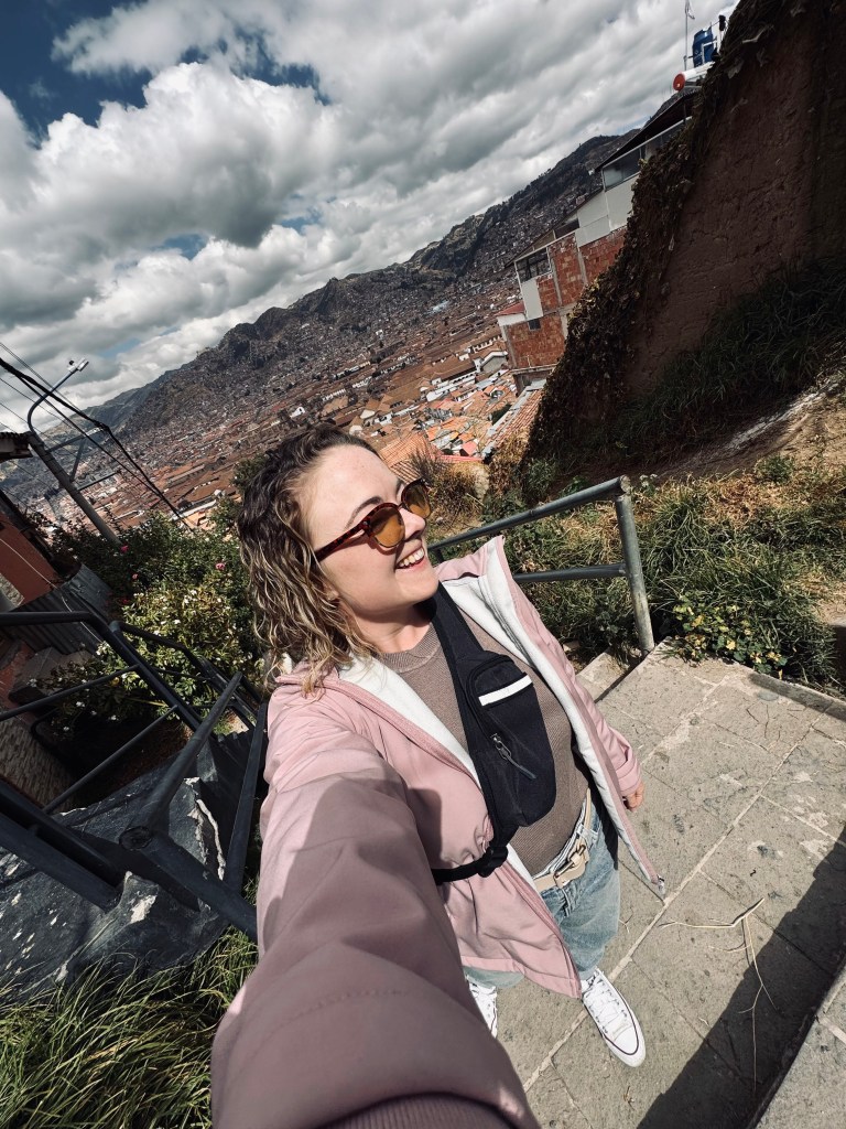 Girl in pink coat taking selfie walking up the stairs with Cusco city in background in Peru