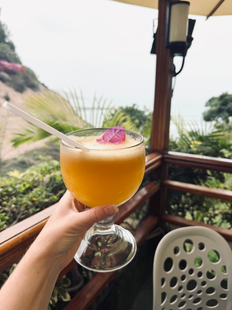Hand holding passionfruit flavored Pisco Sour with ocean view in background in Barranco, Lima, Peru