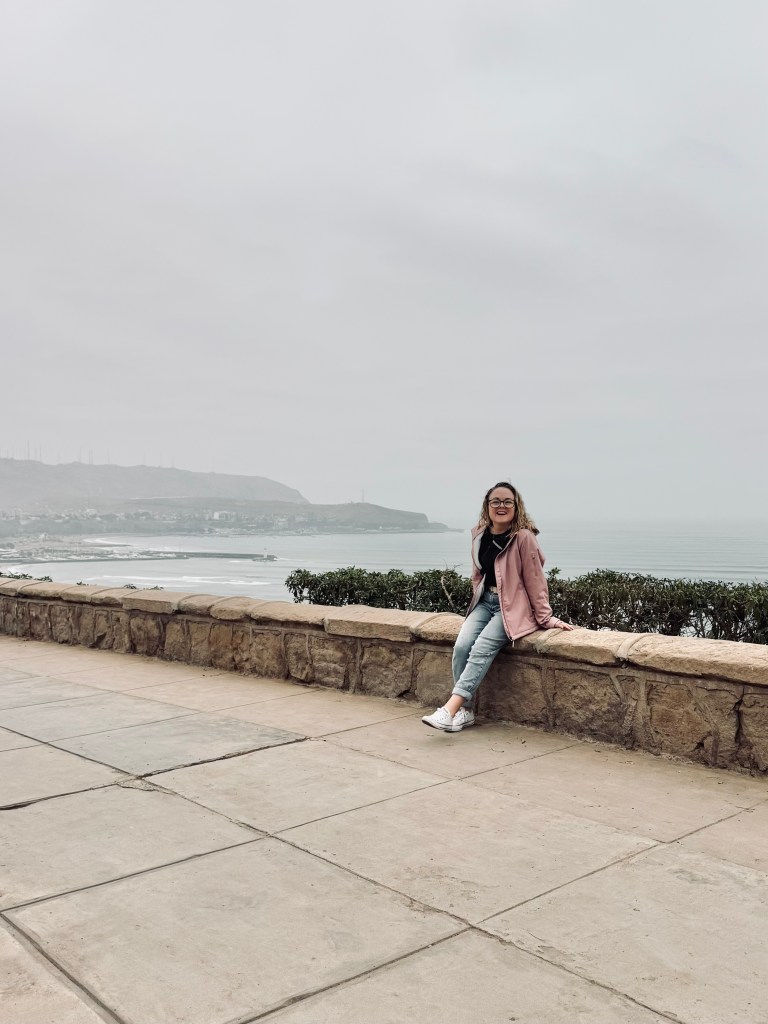 Girl sitting on stone bench with cliffs and ocean and cloudy skies in the background in Miraflores, Lima, Peru