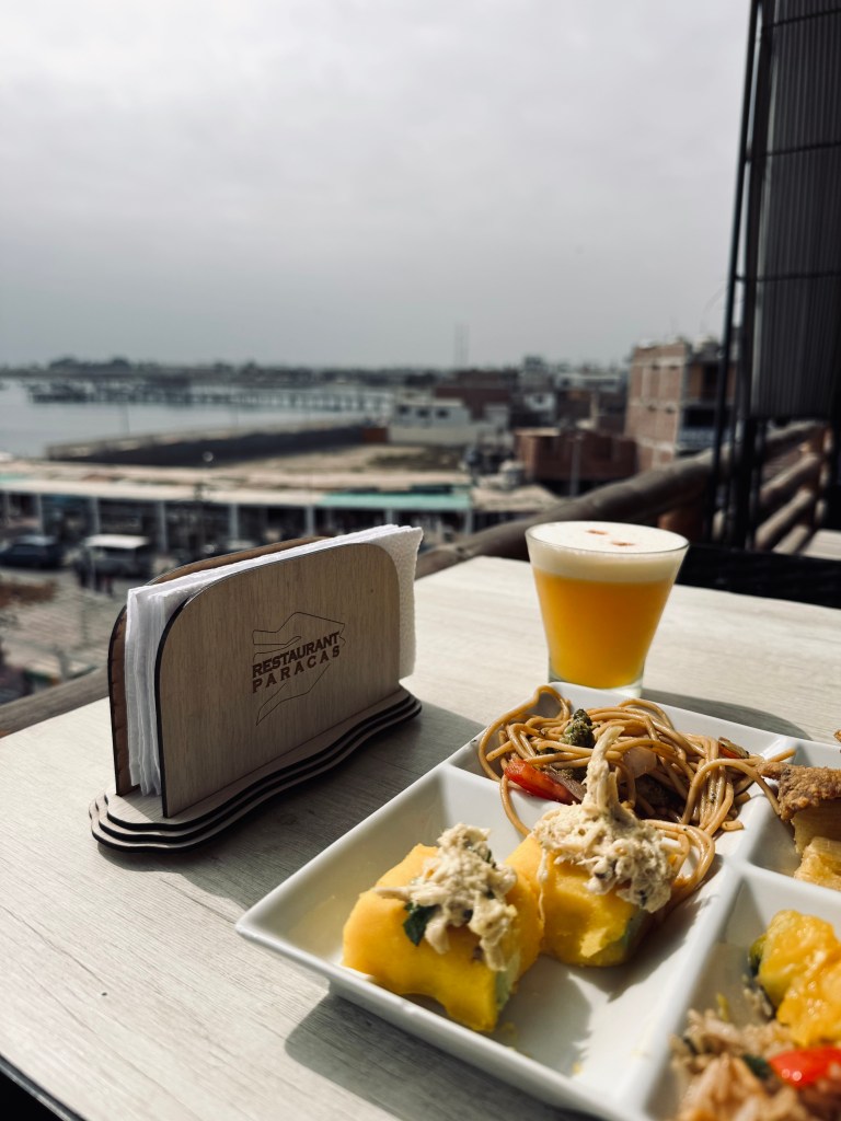 Table with views of ocean in the background. Meal includes causas, noodles and pisco sour in Paracas, Peru.