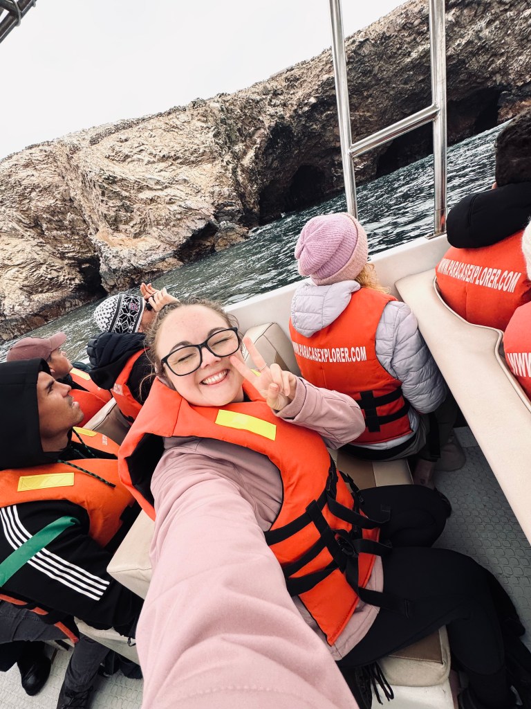 Girl in lifejacket on a boat taking selfie with Islas Ballestas cliffs in Paracas, Peru.