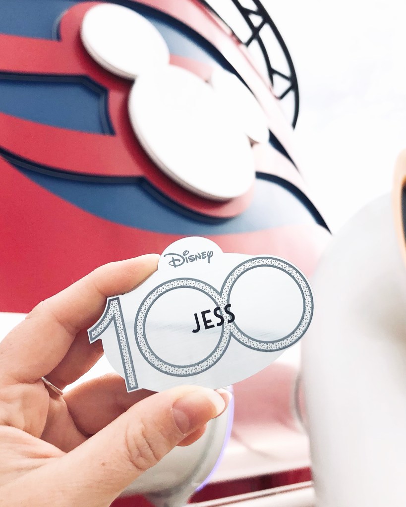 Hand holding Disney 100 cast member nametag with disney cruise line funnels in the background