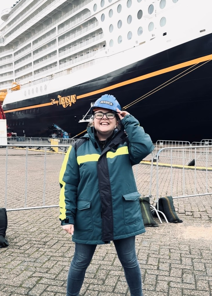 Disney Cruise Line Crew Member smiling with hard hat at the shipyard in front of Disney Treasure Cruise Ship