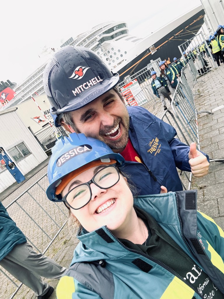 Two disney cruise line crew members wearing hard hats in a shipyard with the Disney Treasure