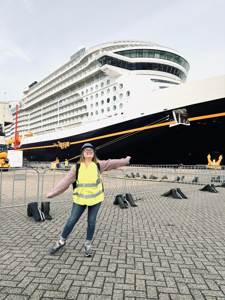 Girl in high vis vest and hard hat smiling at the shipyard with Disney Treasure cruise ship in background