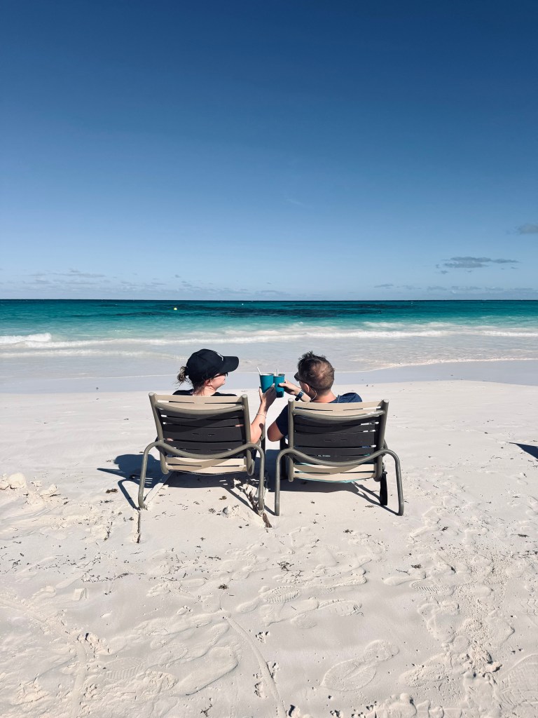 A couple sitting on two beach chairs, cheers-ing a beverage on the beach at Lookout Cay at Lighthouse Point on a Disney Cruise in the Bahamas