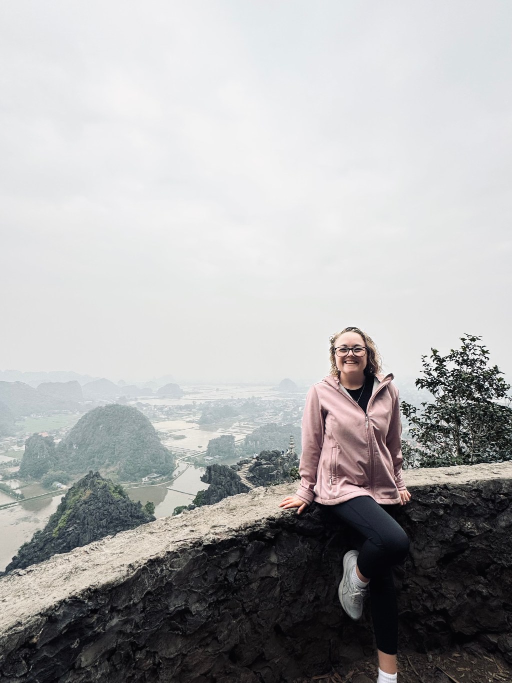 Girl in pink jacket sitting at the top of Mua Cave with view of mountains and rice paddies in distance in Ninh Binh, Vietnam
