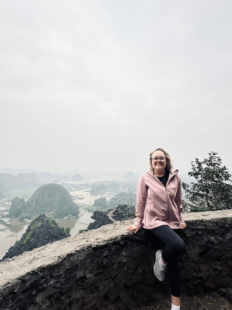 Girl in pink jacket sitting at the top of Mua Cave with view of mountains and rice paddies in distance in Ninh Binh, Vietnam