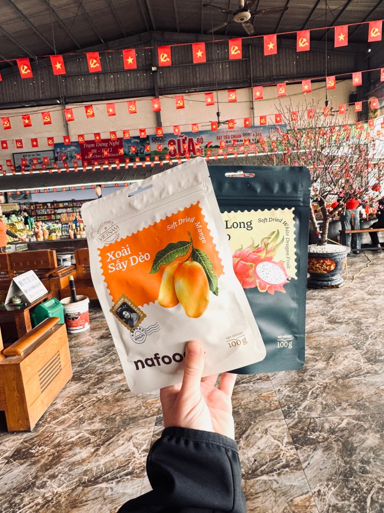 Hand holding dried mango snack with Vietnamese flags in background at a bus rest stop in Vietnam